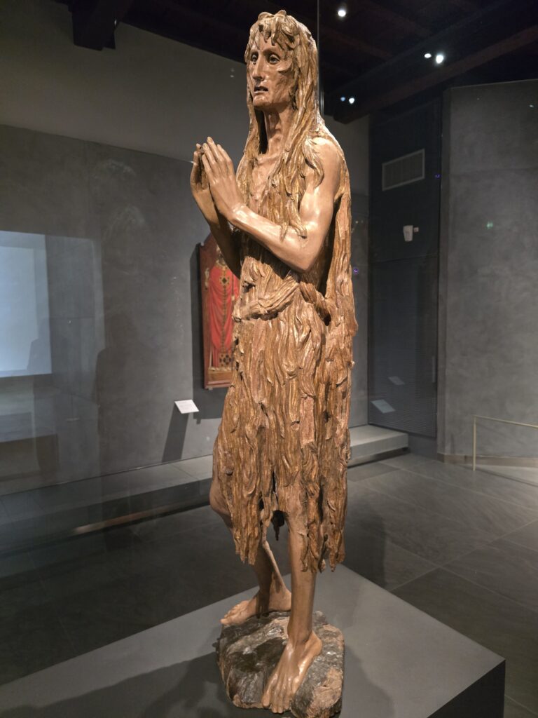 A wooden sculpture of an old woman in ragged clothes with her hands held in prayer